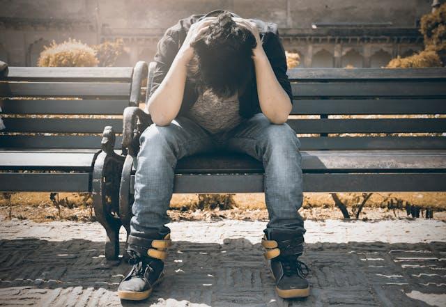 Jeune homme jean et baskets assis sur un banc les mains derrière sa tête baissée