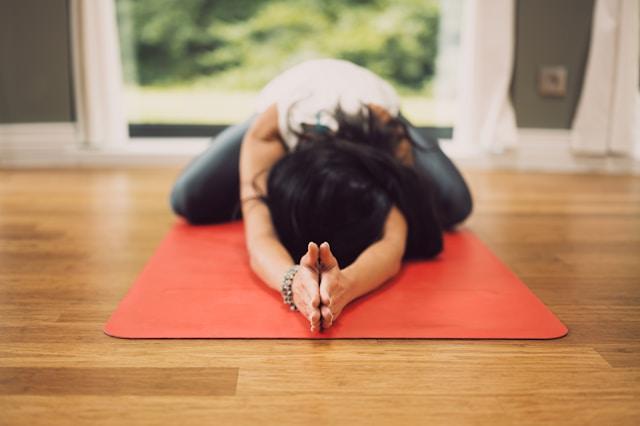 Femme cheveux noirs qui  fait du yoga allongée la tête sur un tapis rouge posé sur parquet avec en arrière-plan fenêtre et jardin