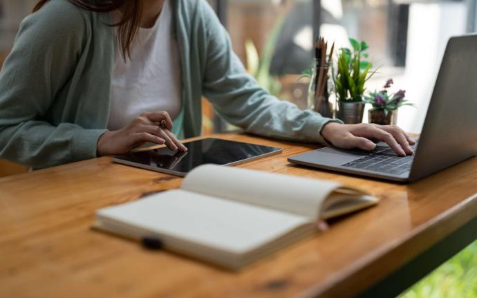 Femme devant ordinateur et tablette avec carnet ouvert au premier plan et plantes sur son bureau en bois