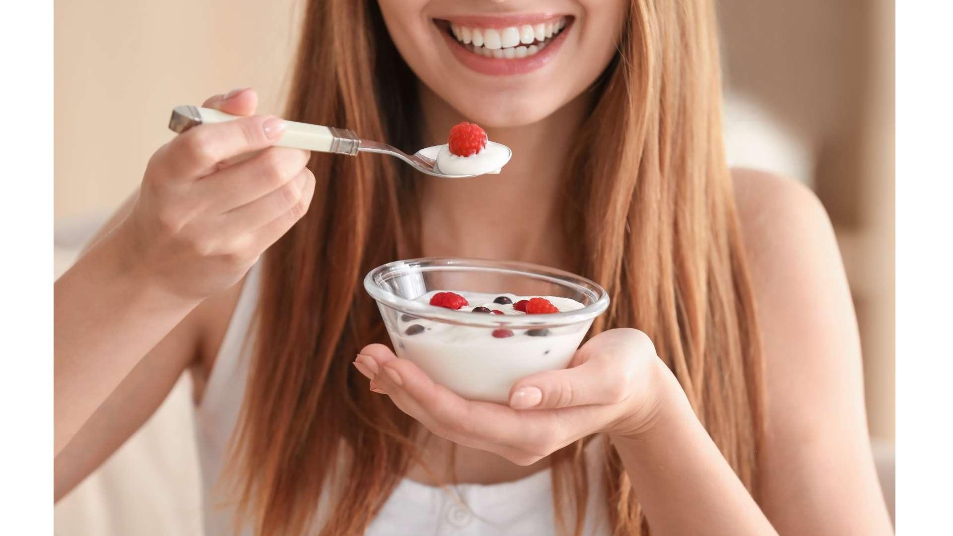 Jeune femme qui sourit en tenant d'une main une cuillère avec yaourt et framboise et de l'autre coupelle avec yaourt et fruits rouges