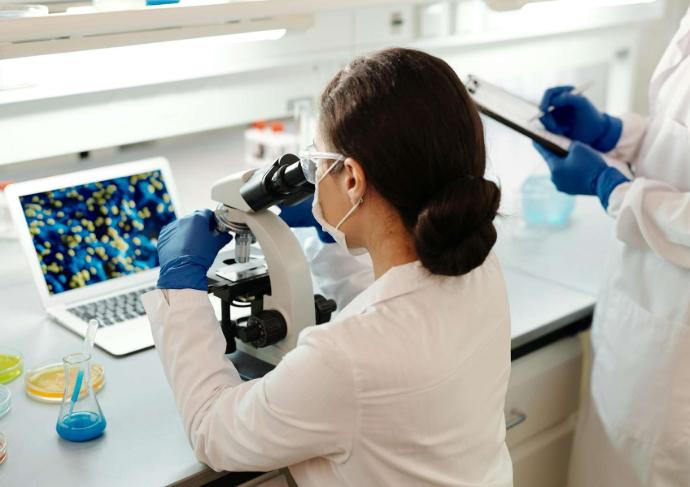 Femme avec blouse et masque blancs et gants bleus sui regarde dans un microscope avec un ordinateur devant elle et une fiole avec liquide bleu à côté