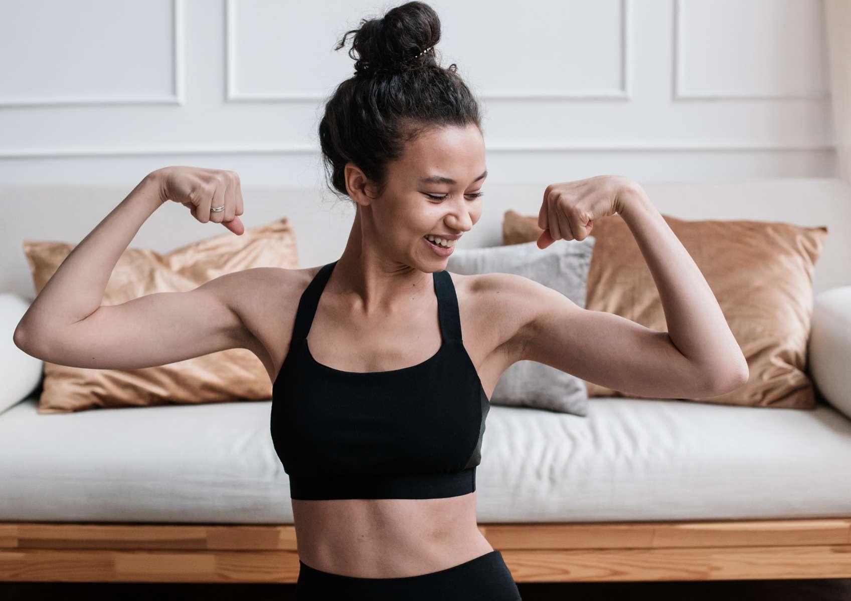 Jeune femme haut et bas noir qui gonfle ses biceps en les regardant en souriant avec canapé en arrière-plan