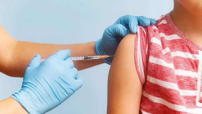 Main avec gant bleu qui tient une aiguille sur le bras d'un enfant avec tee-shirt à rayures blanches et rouges
