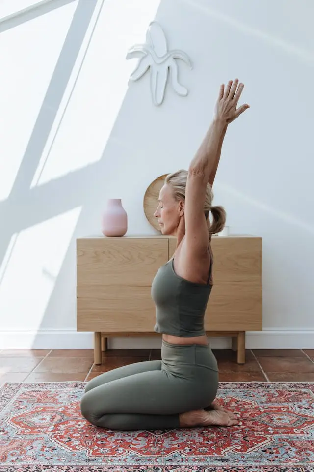 Femme blonde de profil yeux fermés habillée en kaki  assise talons sur les fesses et bras levés avec meuble en bois derrière elle