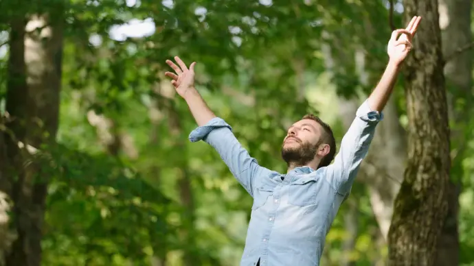Homme avec une chemise en jean bleu clair qui lève les bras en souriant entouré d'arbres