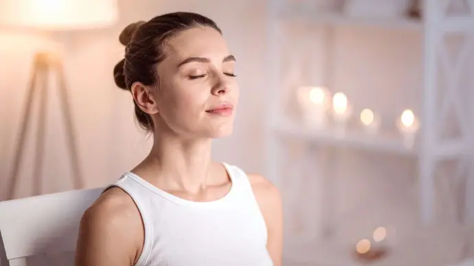 Femme avec débardeur blanc assise sur une chaise blanchel es yeux fermés et visage détendu avec en arrière-plan une lampe et une  étagère blanche avec des bougies