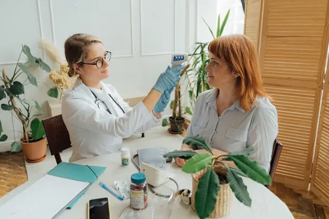 Médecin femme qui prend la température frontale d'une femme assises devant une table ronde blanche avec pochette bleu, plante, téléphone et médicaments en arrière-plan plantes et sur le côté paravent en, bois ccair