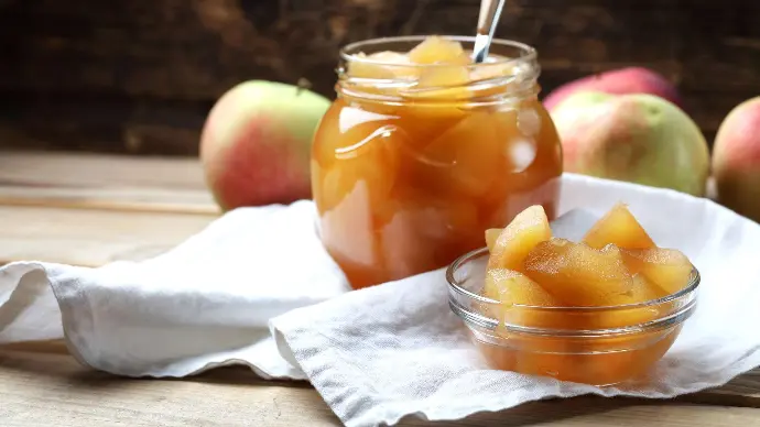 Compote de pommes maison dans une coupelle et un bocal posés sur un torchon blanc sur une table avec en arrière-plan des pommes bicolores rouges et vertes