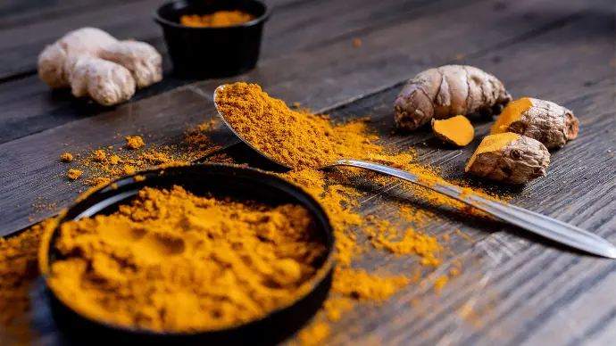 Curcuma en poudre dans coupelle noire,  petite cuillère argentée et racines posés sur table en bois marron foncé